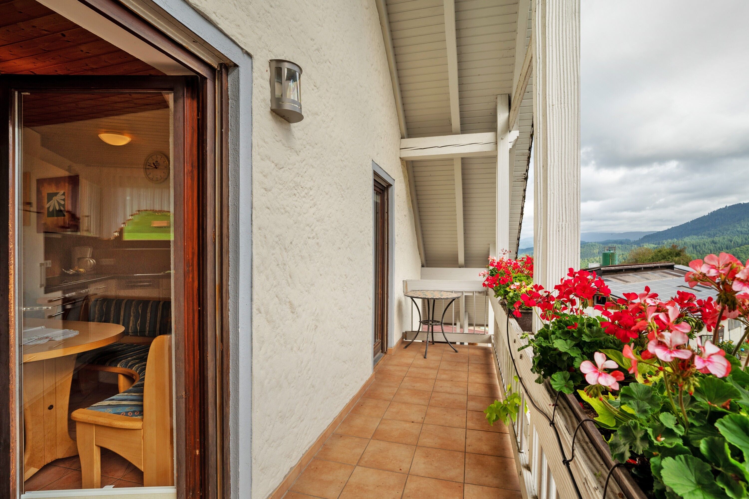 Ein Balkon mit Tisch und Stühlen, Blick ins Grüne und einem Blumenkasten mit roten Blumen.