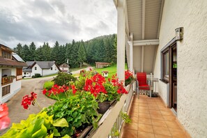 A balcony with potted plants, a red chair, and a view of houses and forest.