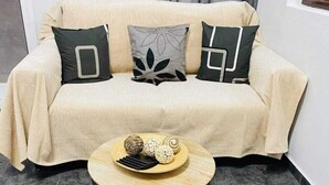 A beige sofa with three black pillows, a round wooden table with a black wireframe base, and decorative items on the table. - Sabbia Calda  apt 8 by Comfortbnb (Perissa)