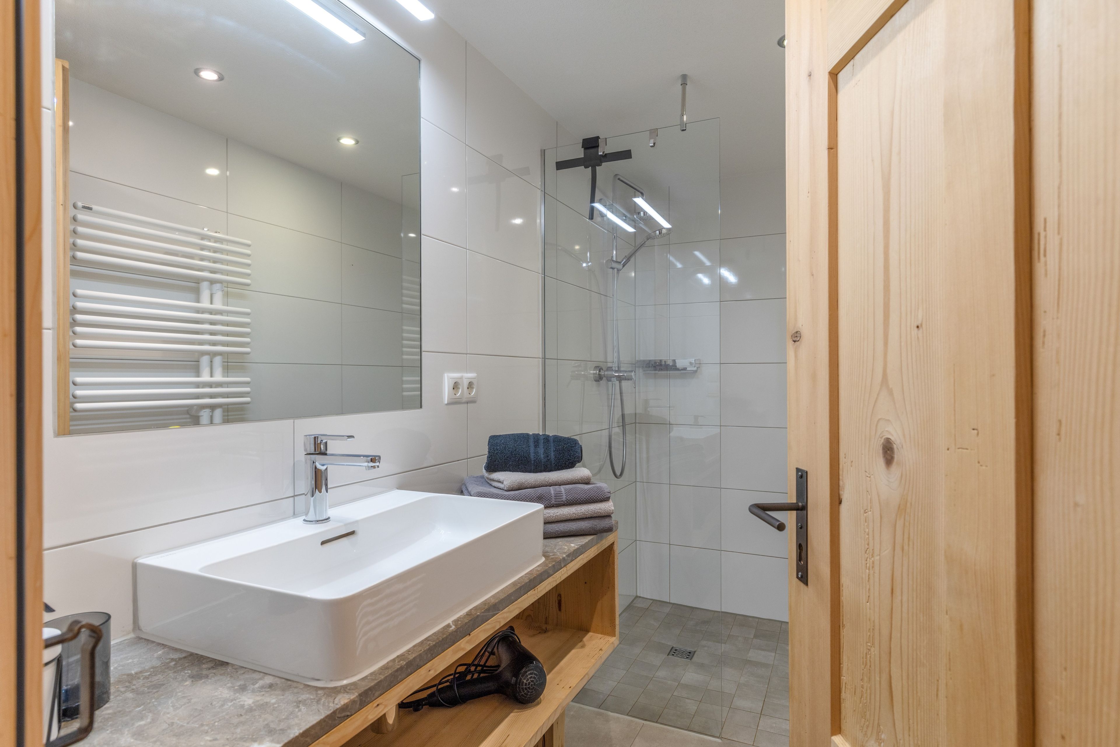 Ein modernes Badezimmer mit großem Spiegel, Waschbecken in Weiß, Dusche und Holztür.
