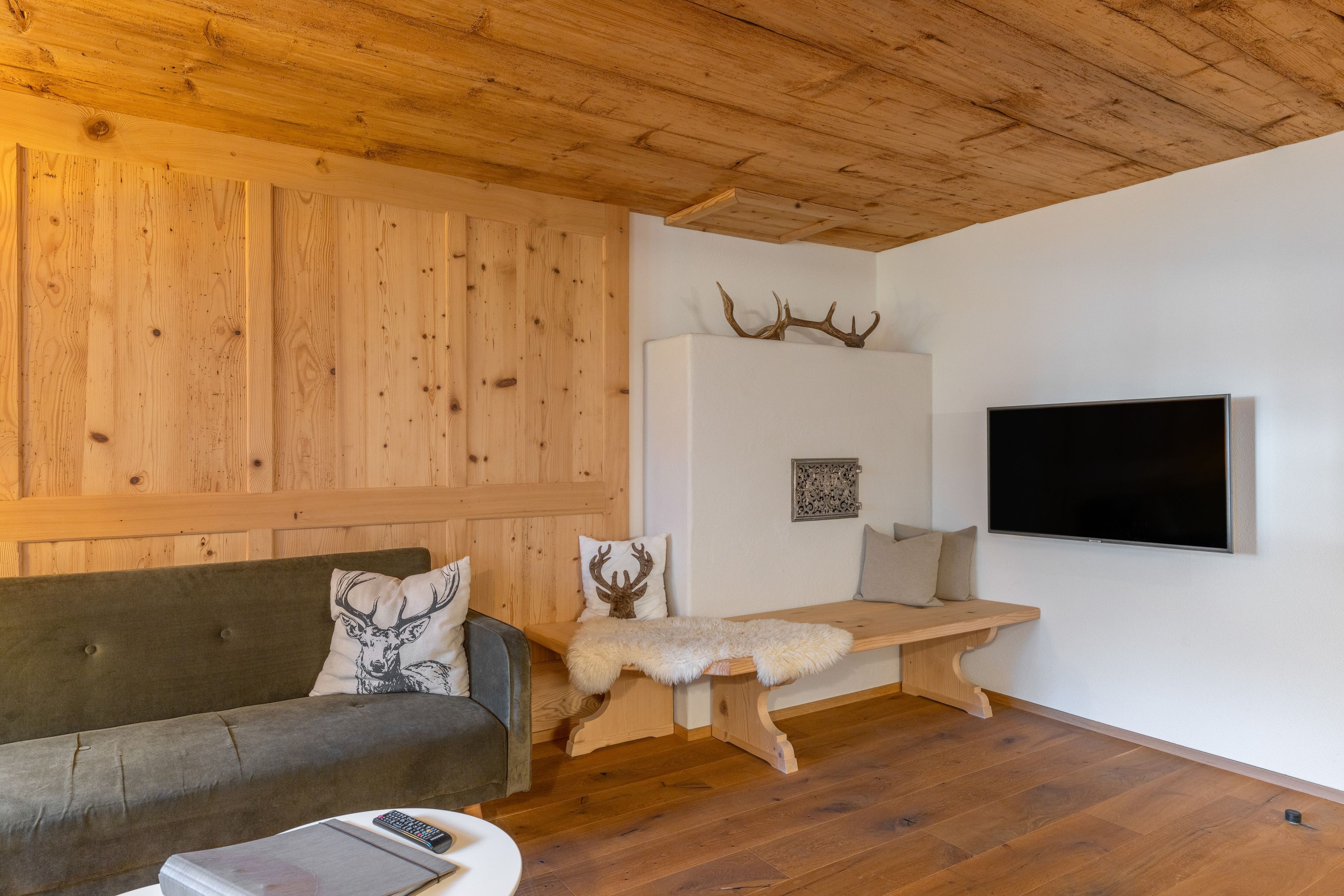 Ein gemütliches Zimmer mit Holzbalkendecke, einem Sofa mit Geweihkissen, einer Holzbank und einem Wand-Fernseher.