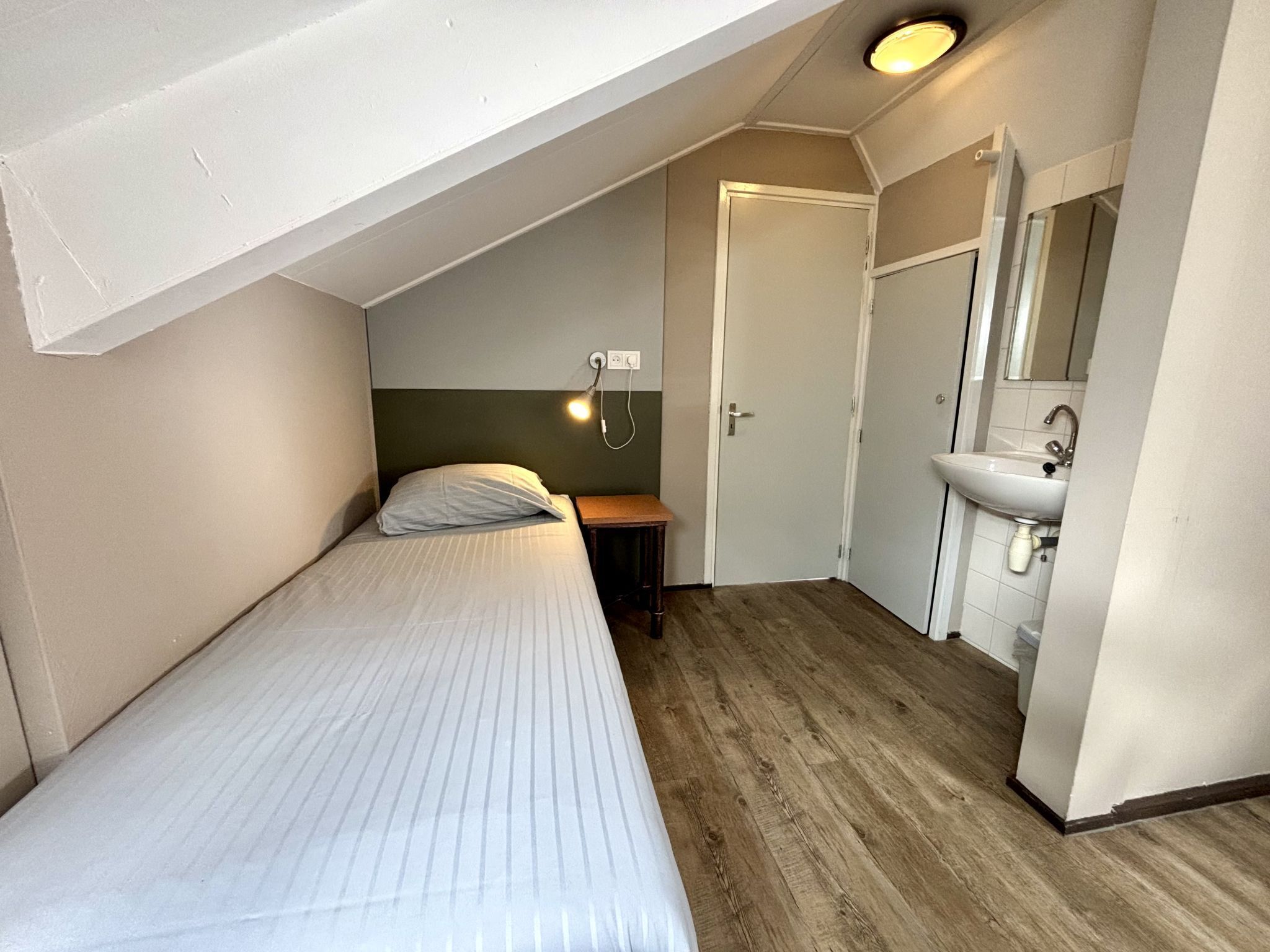 Une petite chambre avec un lit simple, un plafond en pente, une table de chevet, une lampe fixée au mur, une porte et une salle de bain équipée d’un lavabo et d’un miroir.