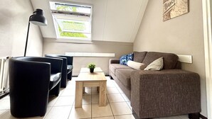 Un salon chaleureux avec un canapé marron, un fauteuil noir, une table basse en bois et un lampadaire noir. Une fenêtre donnant sur un paysage verdoyant se trouve au-dessus du canapé.
