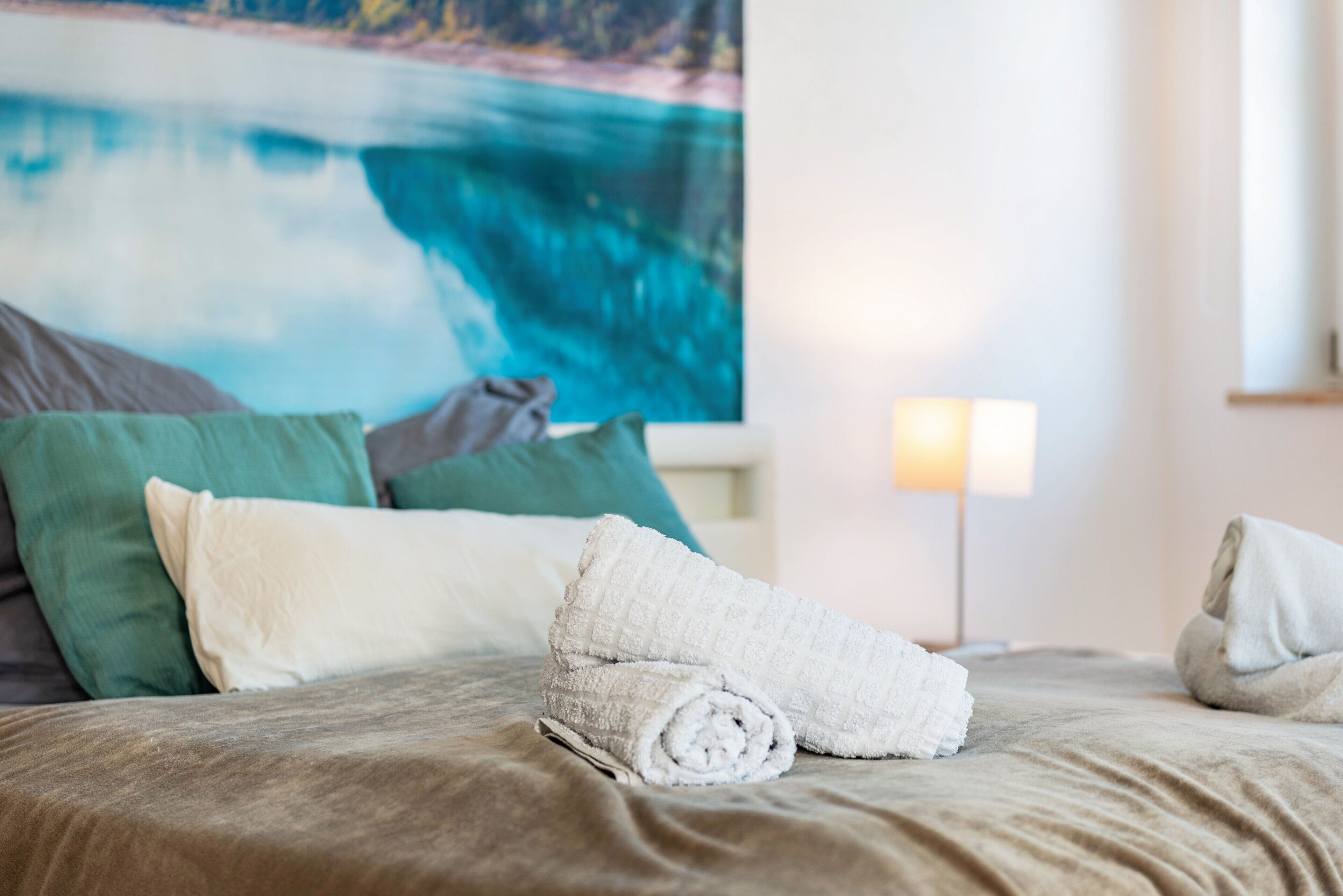 A bed with rolled towels, green and white pillows, and a painting of a coastal scene.