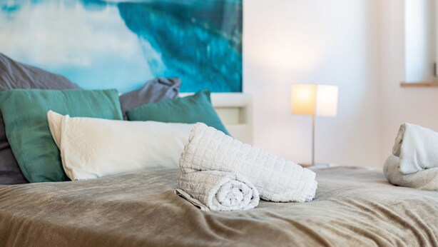 A bed with rolled towels, green and white pillows, and a painting of a coastal scene. - Lake Constance Holiday Home Brunnenberg – Relaxation Close to the Lake (Sipplingen)