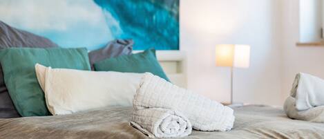 A bed with rolled towels, green and white pillows, and a painting of a coastal scene.