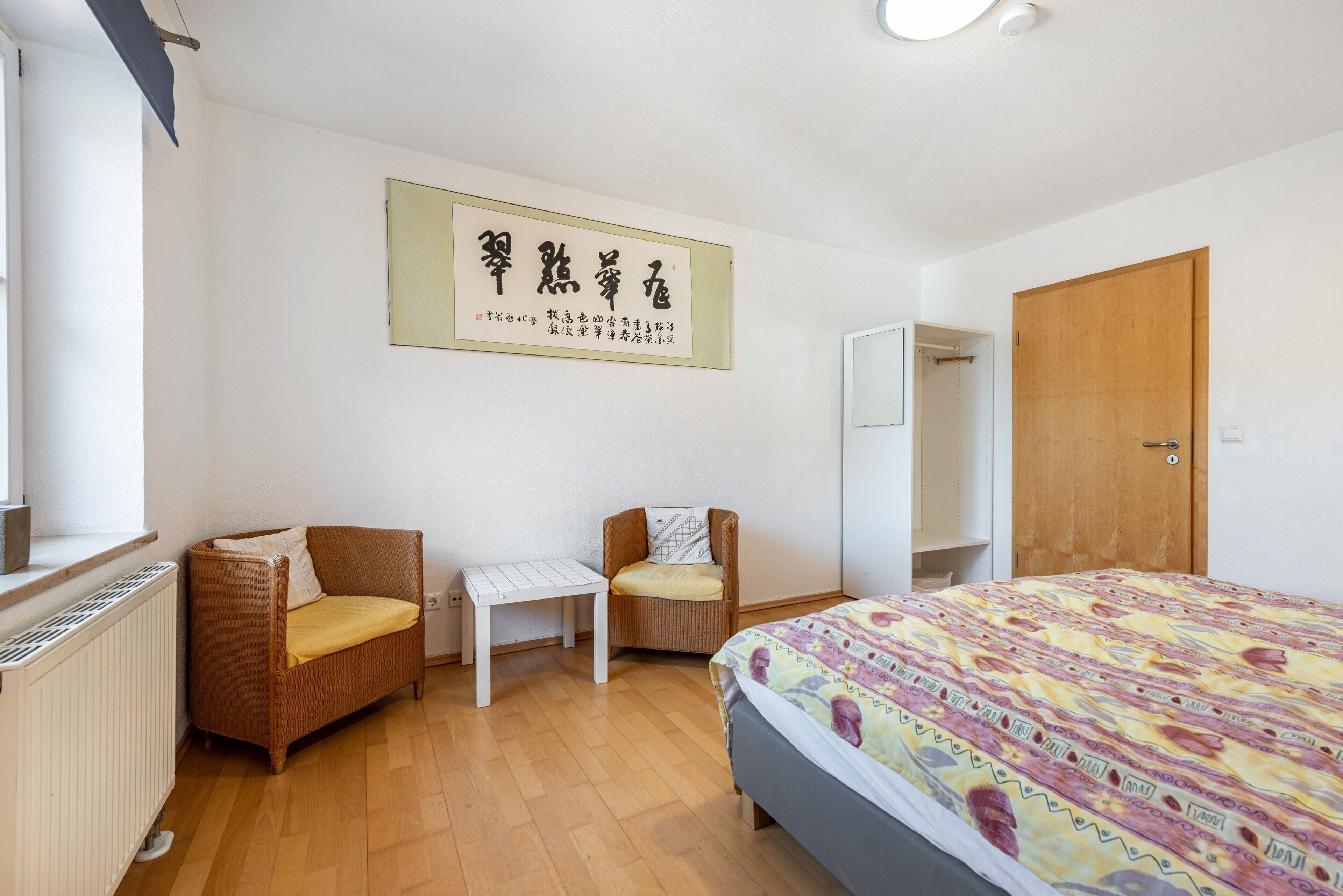 A bedroom with a bed, two chairs, a small table, a radiator, and a wooden door.