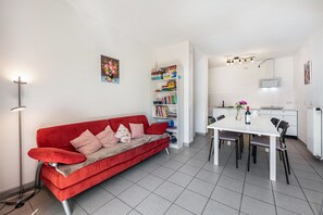 A red sofa with pillows, a white dining table with chairs, a bookshelf, and a floor lamp. - Lake Constance Holiday Home Brunnenberg – Relaxation Close to the Lake (Sipplingen)