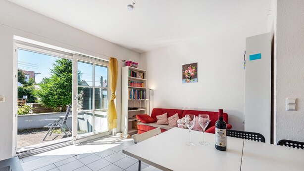 Uma sala de estar moderna com um sofá vermelho, uma mesa de jantar com cadeiras, uma estante de livros e uma janela grande com vista para o exterior.