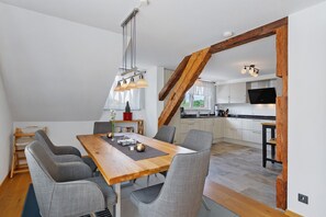 A dining area with a wooden table and grey upholstered chairs, a kitchen with white cabinets and a TV, and a wooden beam structure. - Apartment 'Reiselust' with Private Terrace and Wi-Fi (Stockach)