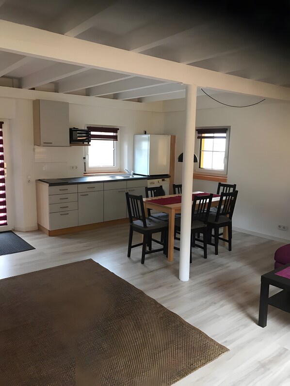 Un comedor con una mesa y sillas, una cocina con armarios y un refrigerador, y una ventana con persianas.
