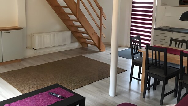 Un salon comprenant un canapé, une table basse, un escalier, un coin repas avec des chaises et un coin cuisine.