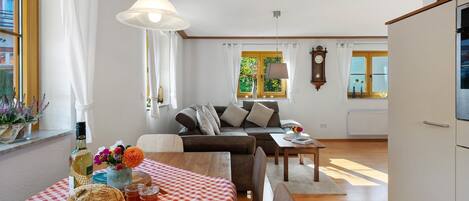 A living room with a sofa, a dining table with a checkered tablecloth, a wooden table with bread and drinks, and a window with curtains.