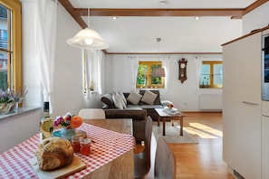 Una sala de estar con un sofá, una mesa de comedor con mantel a cuadros, una mesa de madera con pan y bebidas, y una ventana con cortinas.