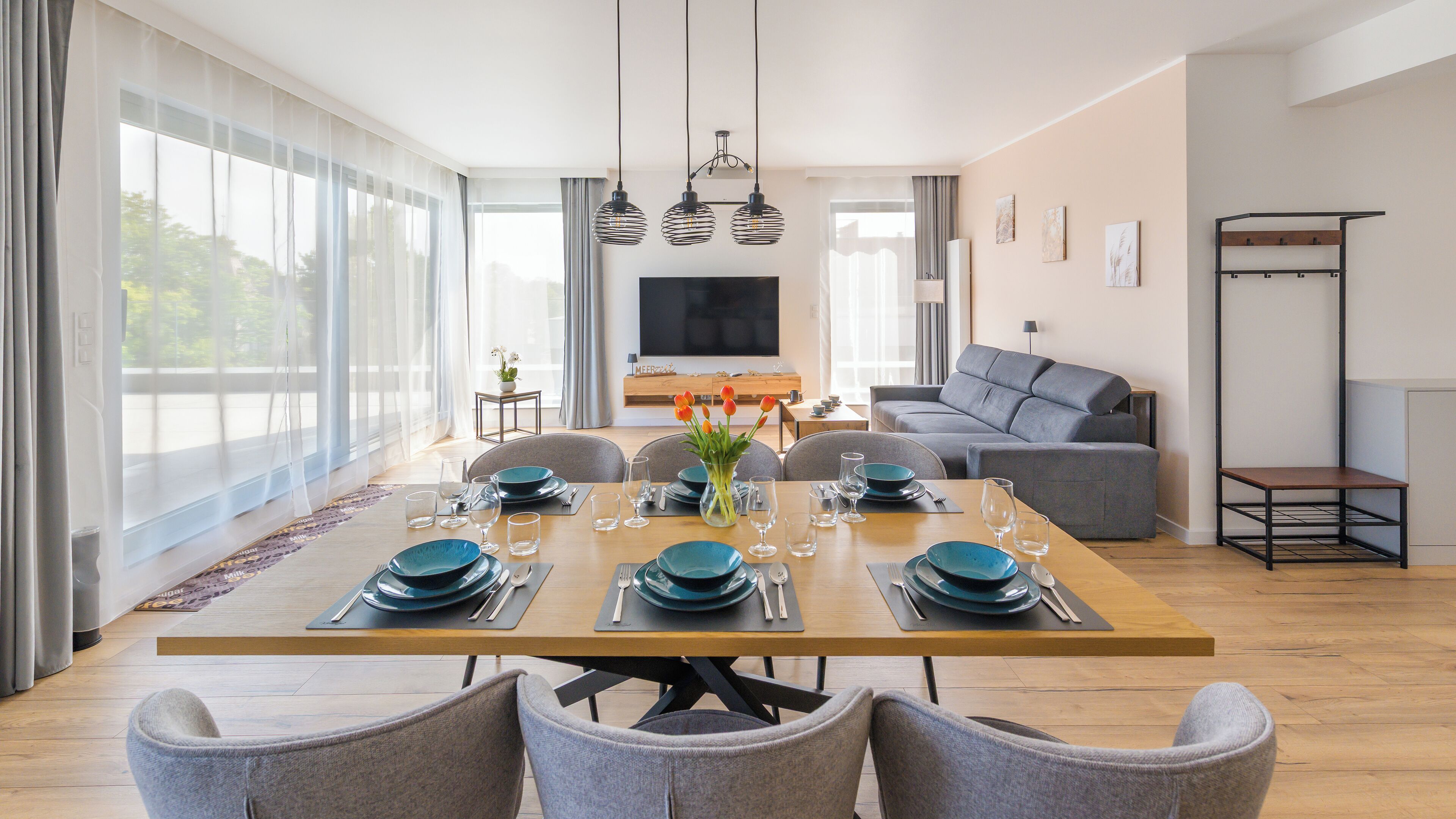 Un comedor moderno con una mesa de madera dispuesta para seis personas, rodeada de sillas grises. En el fondo, se ve un sofá gris y un televisor.