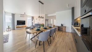 A modern kitchen with a built-in oven, a dining area with a wooden table and grey chairs, and a large window with curtains. - Apartamenty Sun & Snow Sand Dunes (Dziwnów)