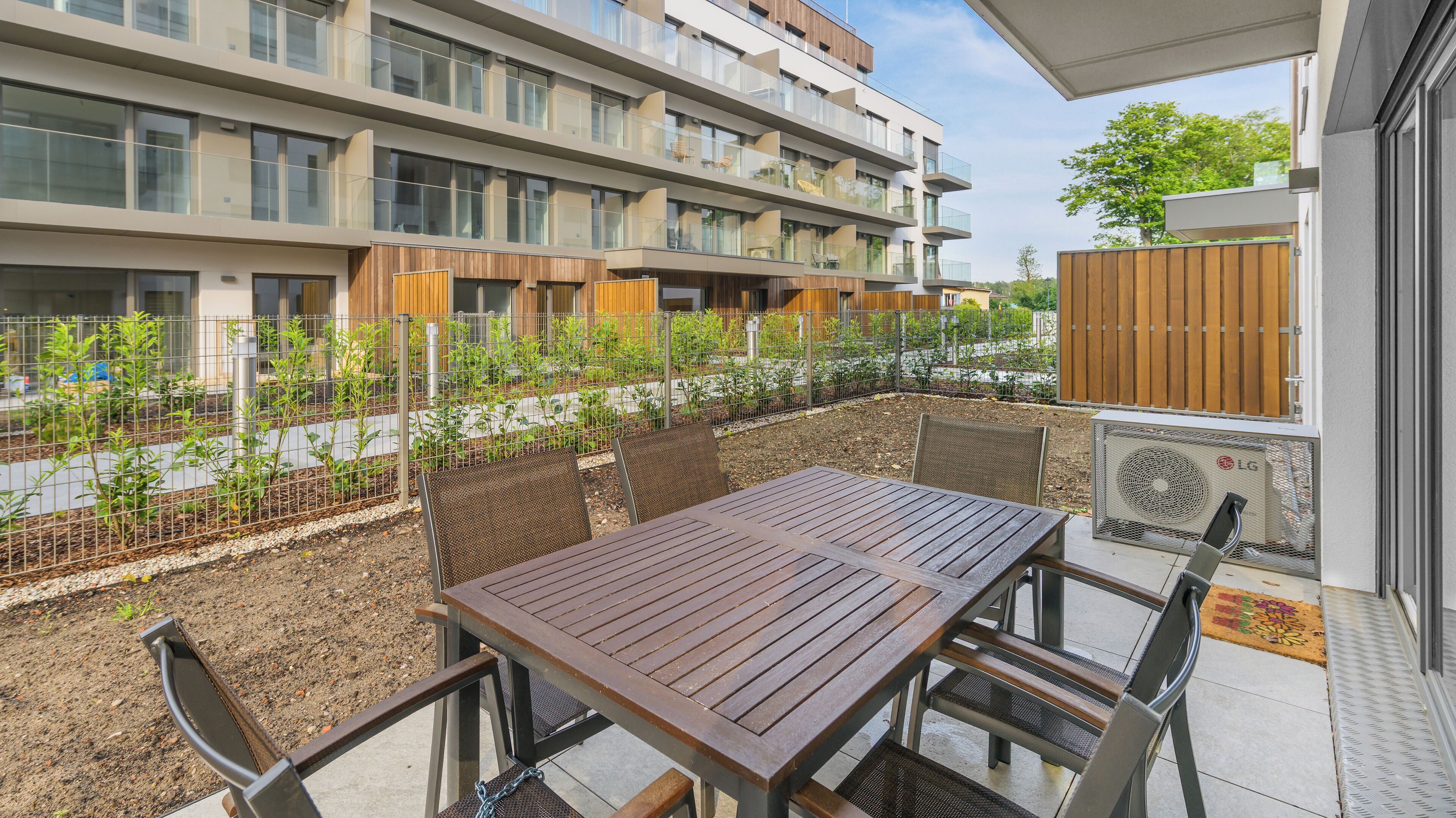 Un balcón con mesa y sillas de madera, una baranda metálica y vistas a un edificio de varios pisos con balcones.