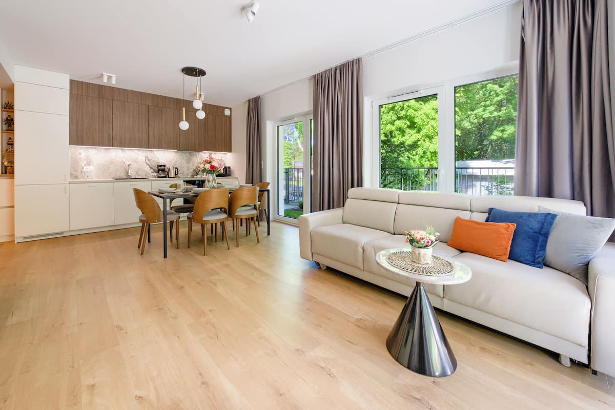 A modern living room with a sofa, dining table, and chairs, a kitchen area, and a large window with curtains.