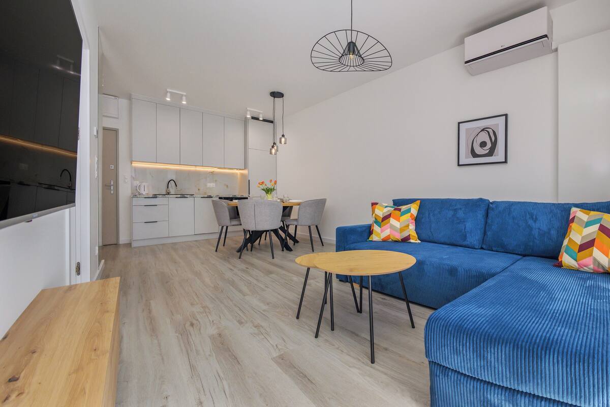 A modern living room with a blue sofa, a wooden coffee table, and a dining area with a kitchen in the background.