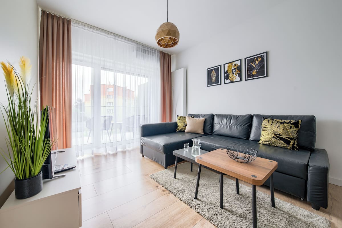 A modern living room with a black leather sofa, a wooden coffee table, and a large window with sheer curtains.