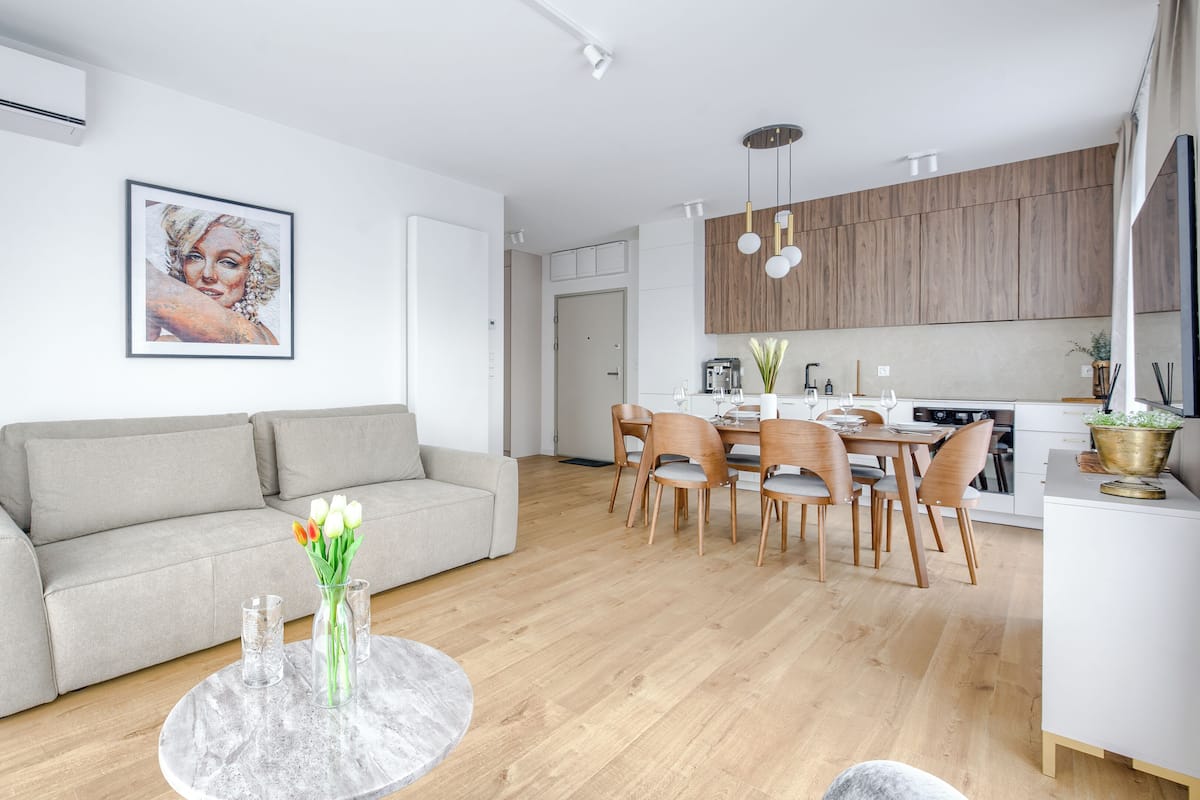 A modern living room with a sofa, dining table, and chairs. There is a kitchen area with wooden cabinets and a stove.