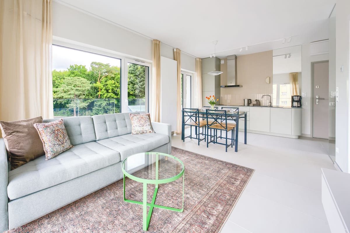 A modern living room with a grey sofa, a glass coffee table, and a view of a balcony with greenery.