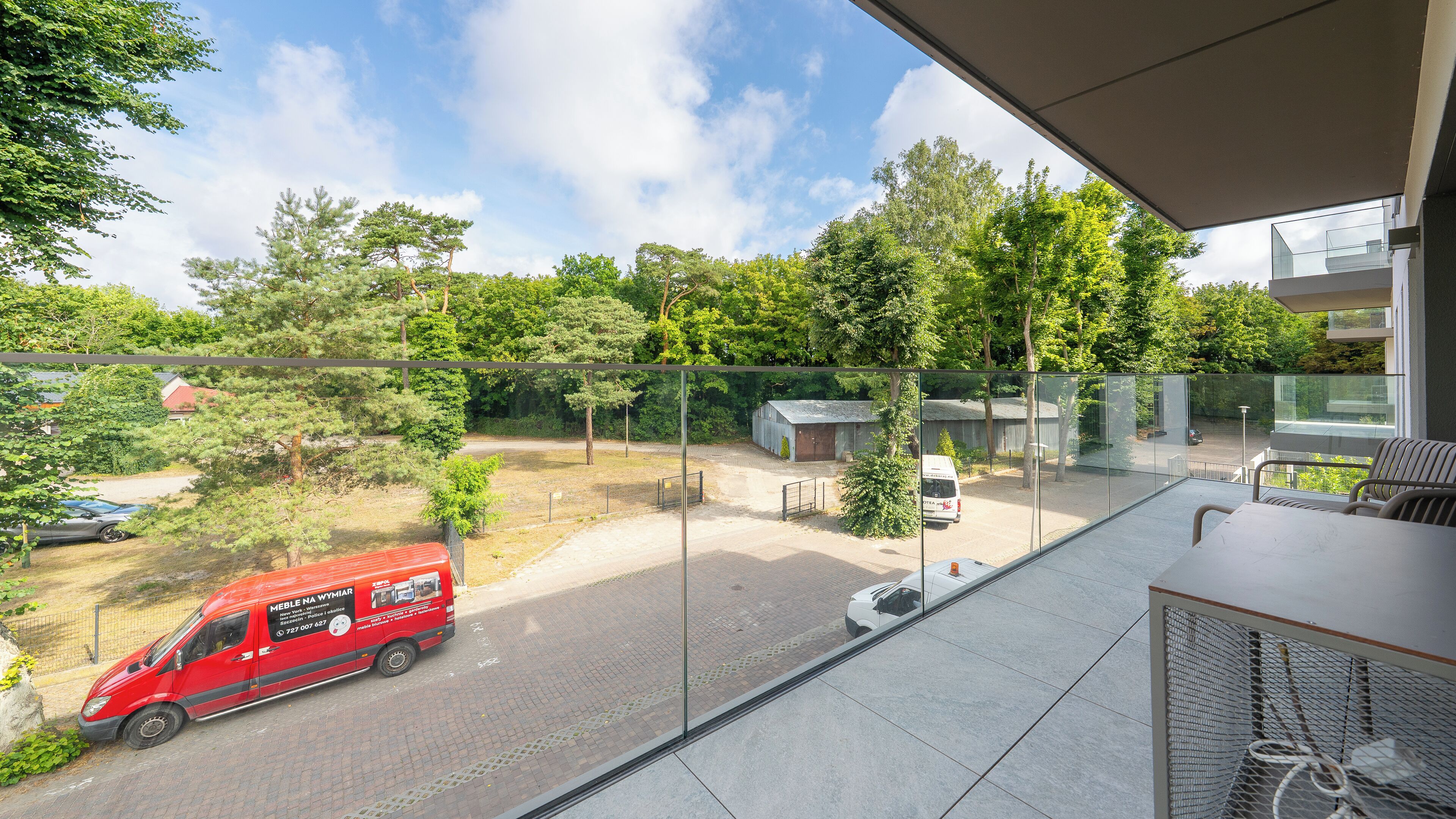 A balcony with a view of a red delivery van on a paved area, surrounded by trees and a building with a glass facade.