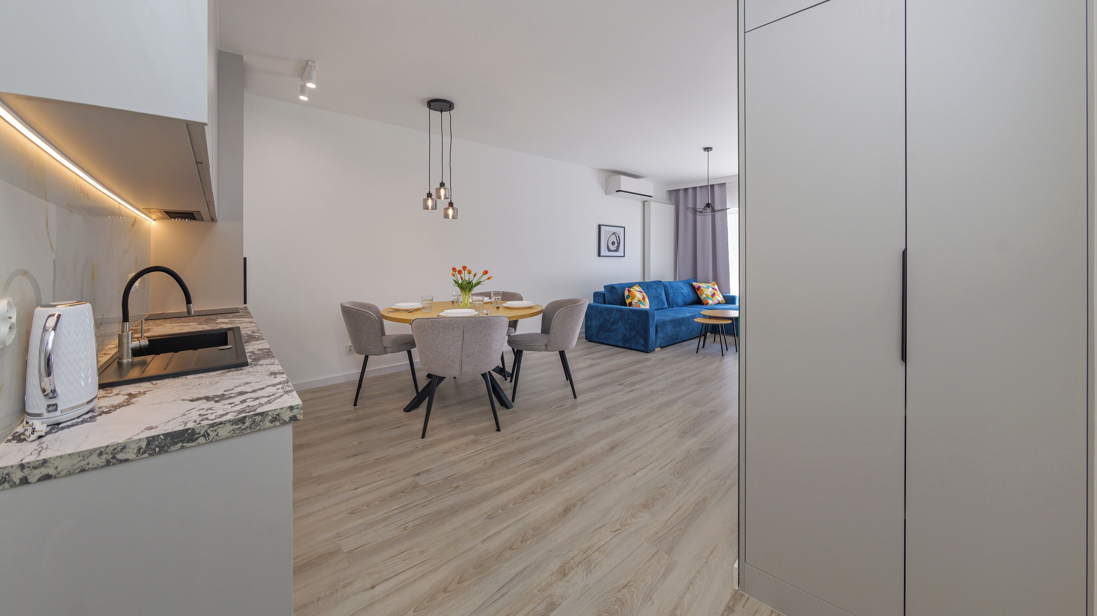 Een moderne keuken met een marmeren aanrecht, een eethoek met een ronde tafel en stoelen, een blauwe bank en een kleine tafel met een bloevas.