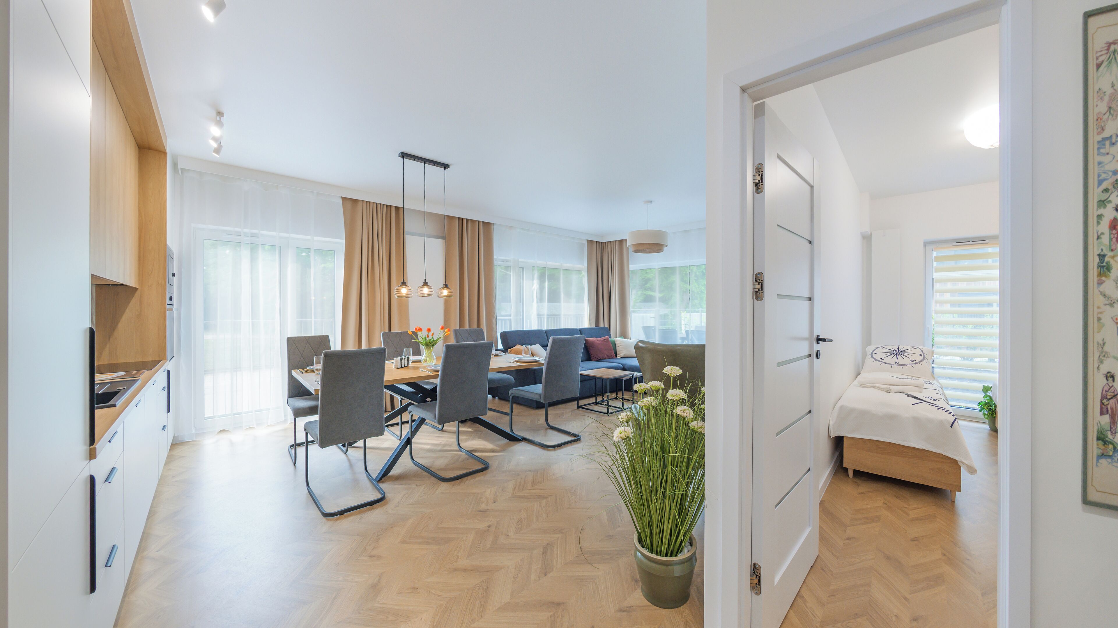 A modern living room with a sofa, dining table, and chairs, a kitchen area, and a bedroom with a bed and window.