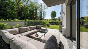 Un espace de vie extérieur moderne, doté d’un canapé d’angle, d’une table basse en bois et offrant une vue sur un jardin luxuriant et les bâtiments environnants.