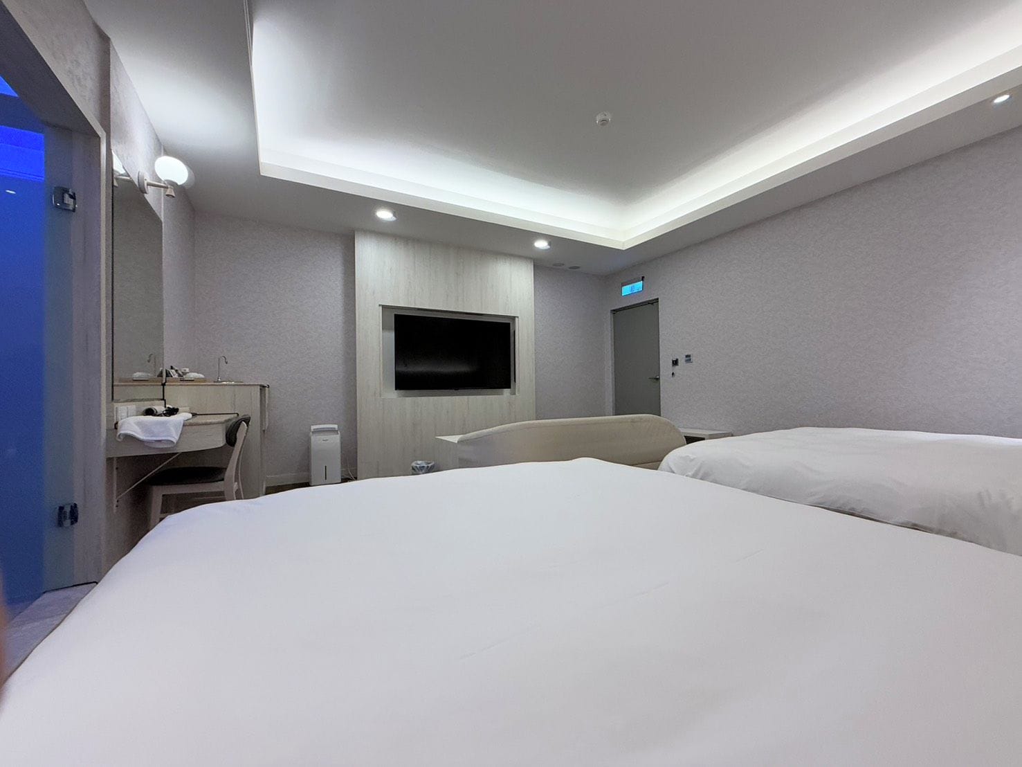 Una camera d'albergo con un letto, una scrivania con una sedia, una TV fissata al muro e una porta con il segnale di uscita.