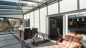 Eine überdachte Terrasse mit einer Liege, einem kleinen Tisch und einem Korbsessel.
