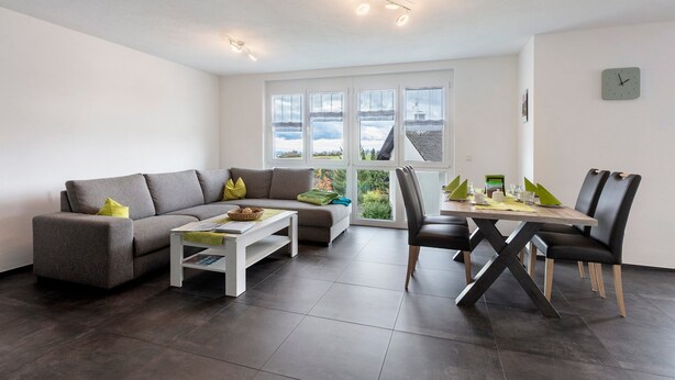 Un salon moderne comprenant un canapé, une table basse, une table à manger avec des chaises, de grandes fenêtres et une horloge au mur.