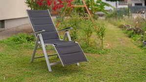 Una sedia da giardino con struttura metallica e seduta nera, posizionata su un prato verde con una casa e piante sullo sfondo.