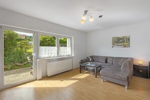 Un salon moderne avec un canapé gris, une table basse et une vue sur un jardin grâce à de grandes fenêtres.