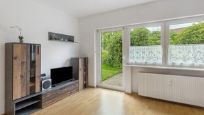 Ein modernes Wohnzimmer mit Flachbildfernseher, Holzschrank und großem Fenster mit Blick ins Grüne.