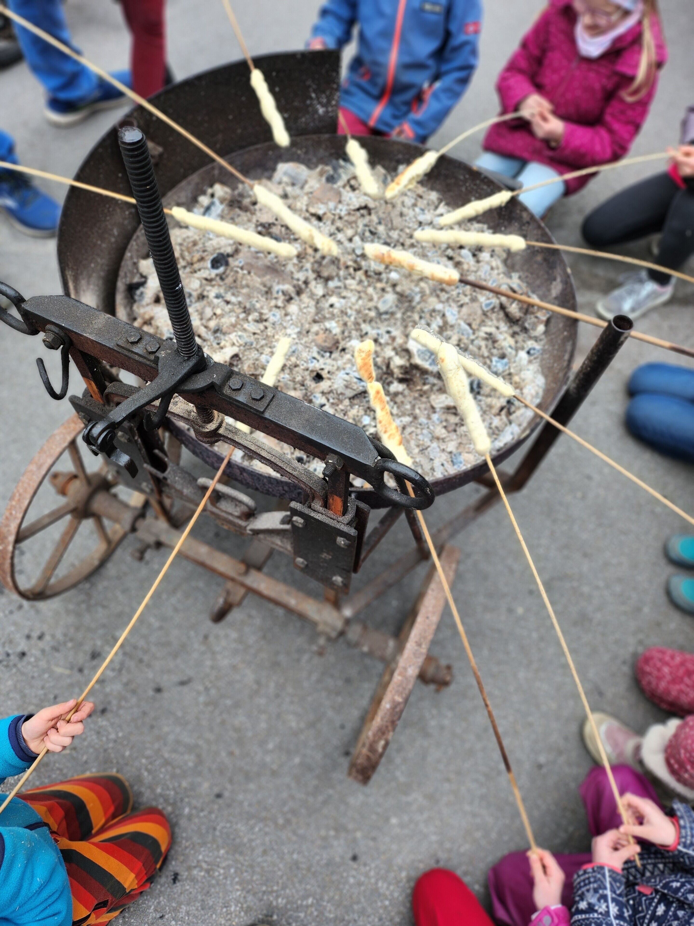 A traditional cooking setup with a large metal pot and a metal rack for skewers, surrounded by people.