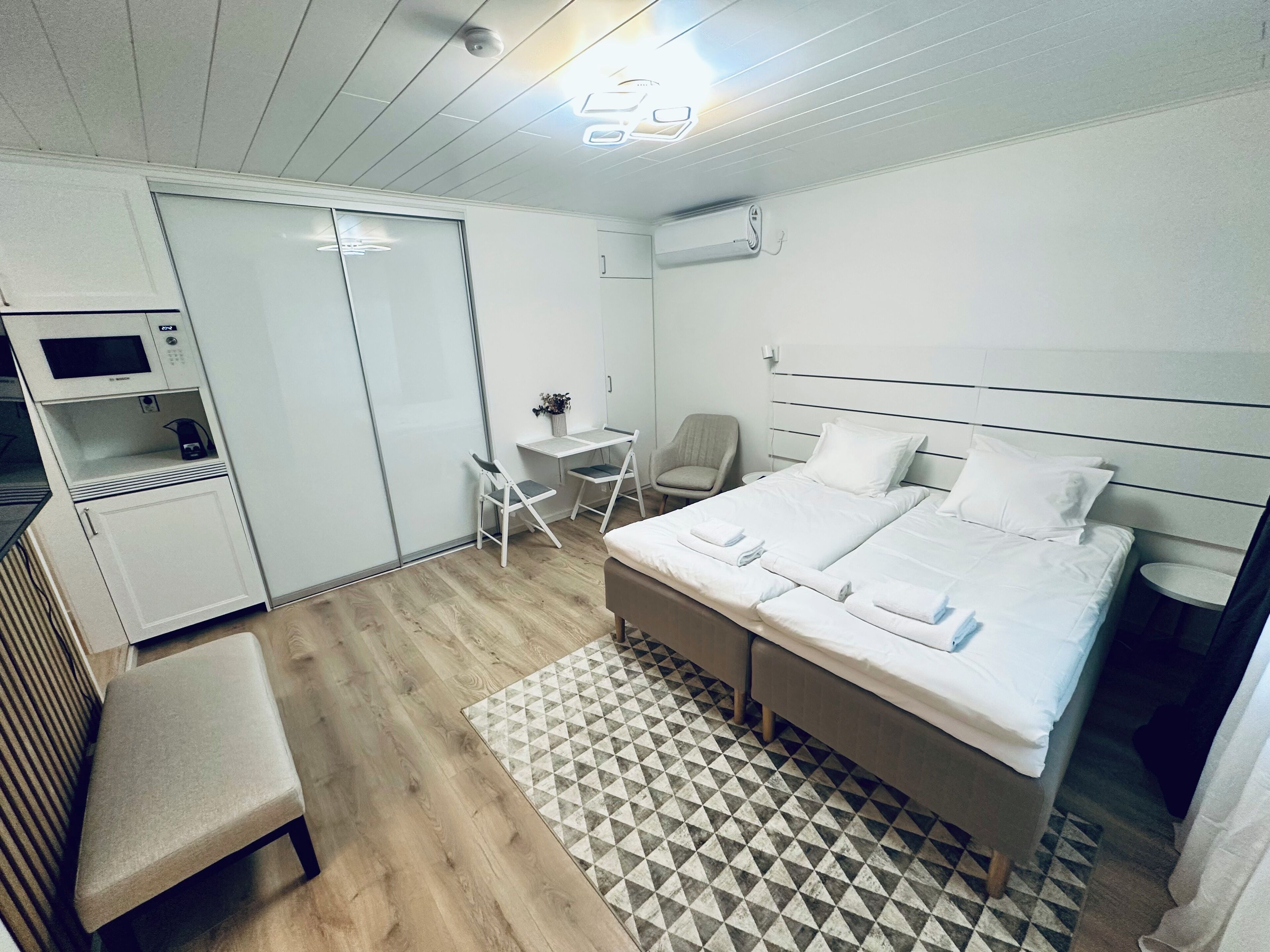 A modern hotel room with a bed, a desk, a chair, a microwave, and a small table.