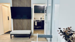 A modern living room with a sofa, a flat-screen TV mounted on a wooden panel, a kitchen area with dark cabinets and a white refrigerator, and a small table with a vase of flowers. - Katariina Apartments (Kotka)