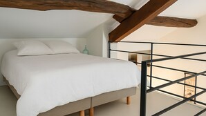 Ein Schlafzimmer mit Bett, Holzbalken, Blick ins Freie und einem kleinen Tisch mit einer Flasche.