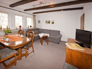 Uma sala de jantar com uma mesa e cadeiras de madeira, um sofá cinza, uma televisão e uma janela com cortinas.