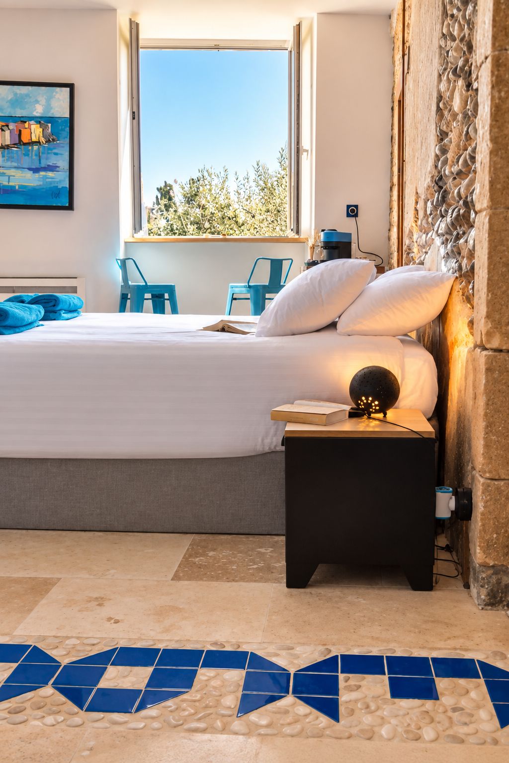 Une chambre à coucher avec un lit, une table de chevet, une fenêtre donnant sur un beau paysage, un tableau au mur et un sol carrelé.