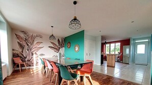 Un vaste espace dédié à la salle à manger, avec une longue table et des chaises aux couleurs vives, une cuisine en arrière-plan et une grande plante sur le côté gauche.