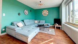 Un salon avec un canapé d’angle, une table basse et un téléviseur. Les murs sont décorés et il y a une fenêtre avec vue.