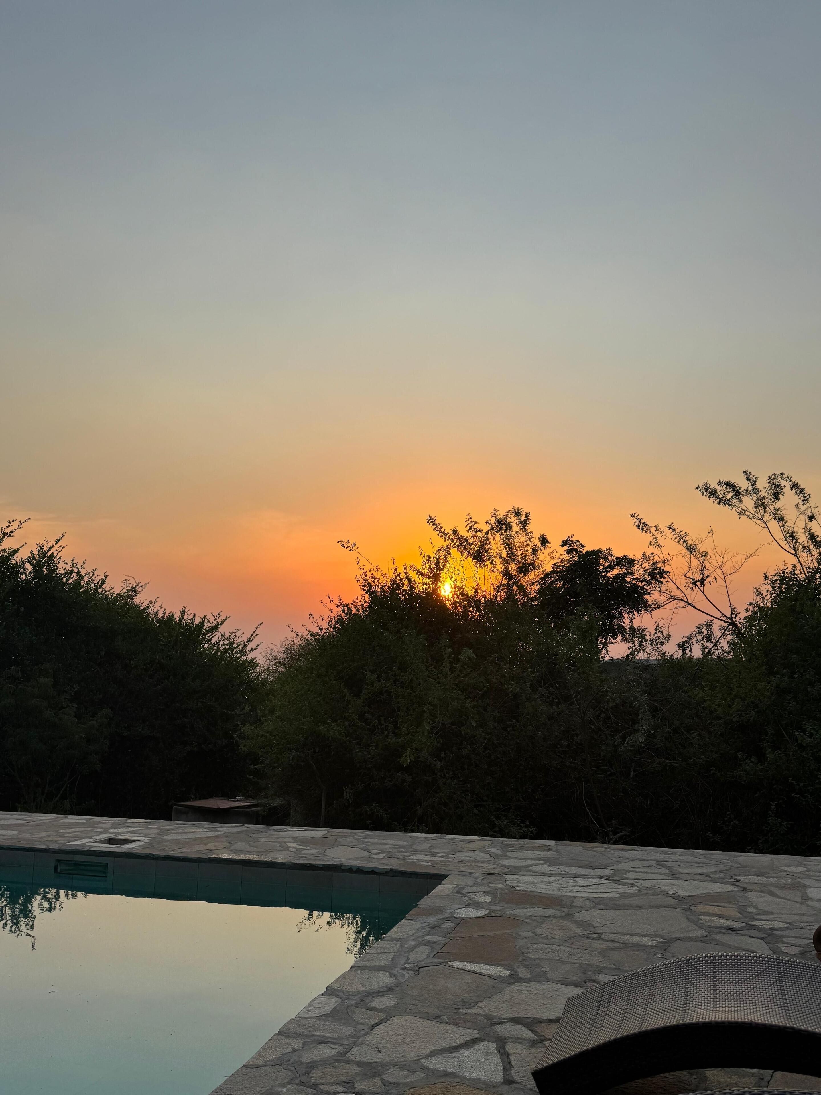 Une piscine en pierre offrant une vue dégagée sur le coucher de soleil, avec en arrière-plan un espace paysager agrémenté d’arbres.