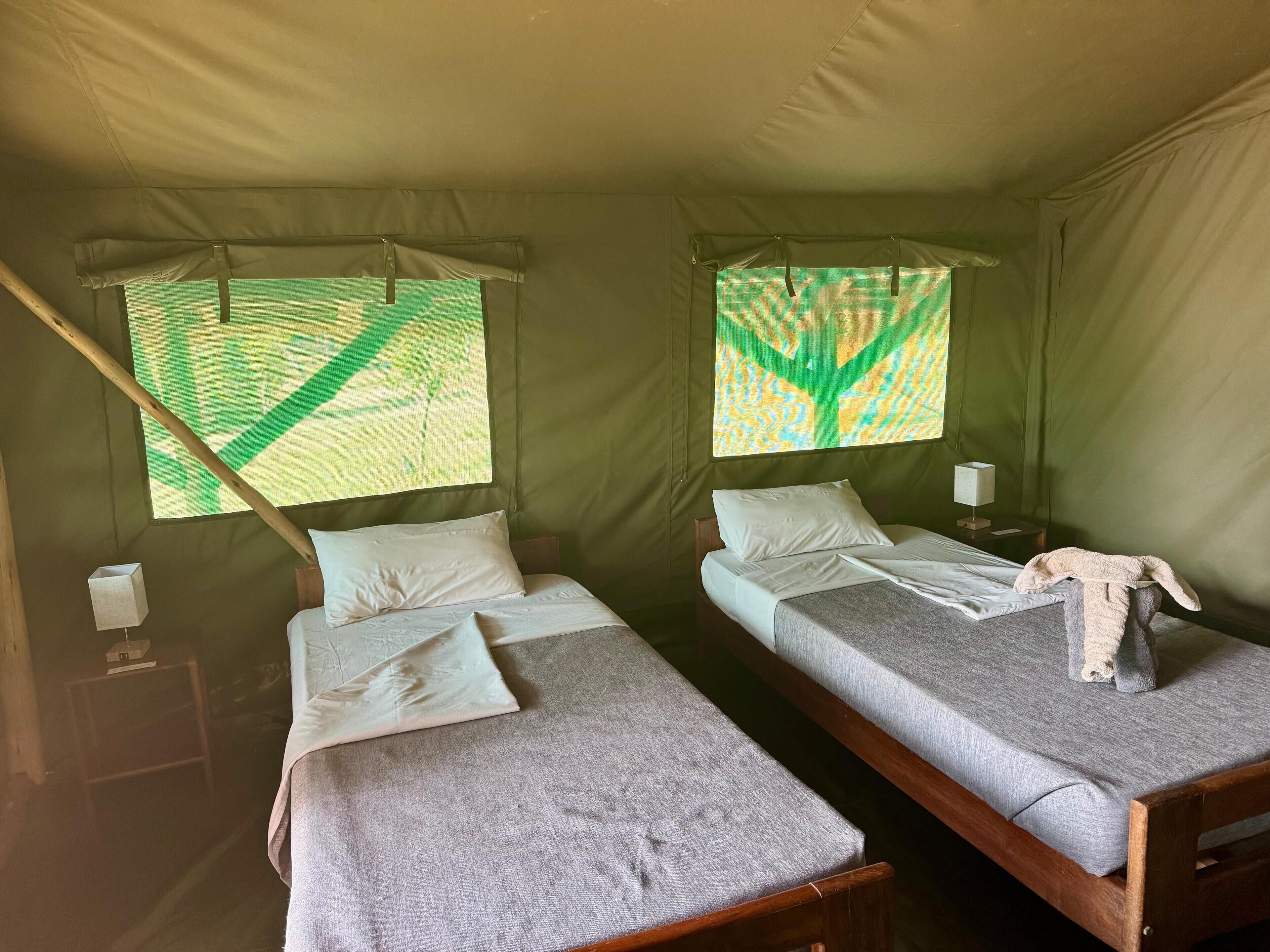 A tent with two beds, each with pillows and a blanket. There is a small table with a lamp and a box on it.