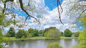 A serene lakeside view with a house in the background, surrounded by lush greenery and trees. - Cortland Riverfront Retreat (Cortland)