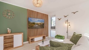 Ein modernes Wohnzimmer mit einer Wandpaneelaus Holz, einer Wanduhr, einem Flachbildfernseher, einem Sofa mit grünen Kissen und einem kleinen Tisch mit einer Flasche und Gläsern.