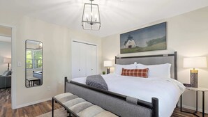 A neatly arranged bedroom with a bed, bedside lamps, a bench, a mirror, and a painting on the wall. - The Rover - Market District - 1m to DT Bentonville (Bentonville)