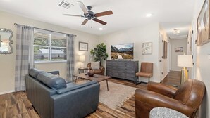A modern living room with a sofa, armchair, and coffee table, a ceiling fan, and a view of the outdoors through large windows. - The Rover - Market District - 1m to DT Bentonville (Bentonville)